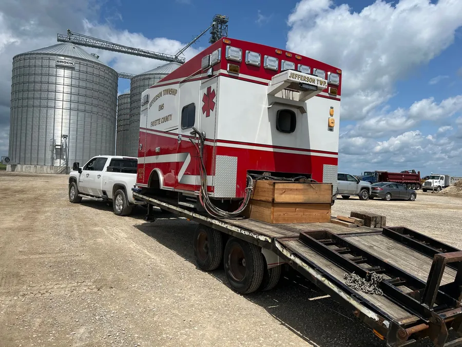 A red and white ambulance box, lifted clear of its chassis and set on timber blocking on a flatbed trailer behind a pickup, with grain bins in the background.