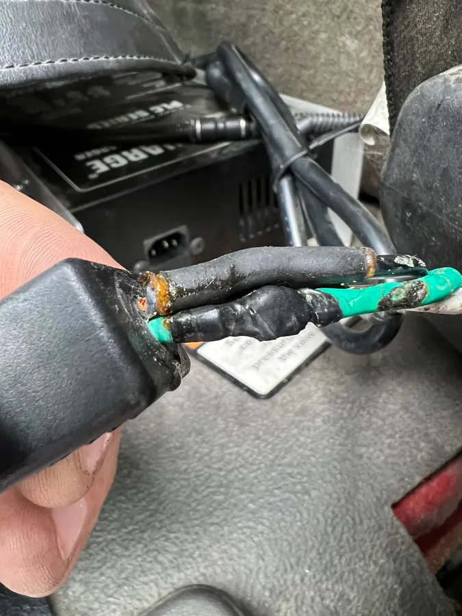 A hand holding a damaged plug from an onboard battery charger, its black and green ground wires crushed and corroded where they leave the moulded connector.