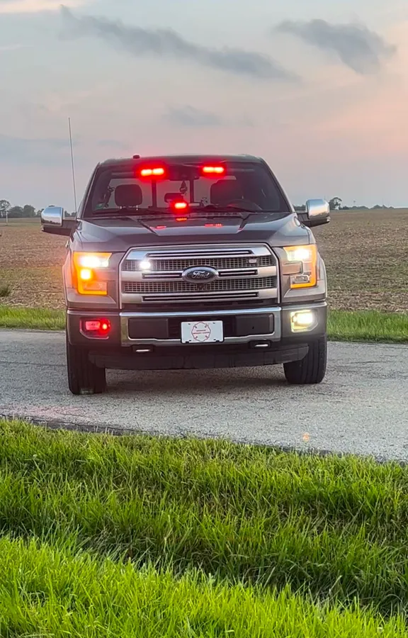A Ford F-150 seen head-on at dusk, red warning lights lit across the windscreen header and behind the grille.