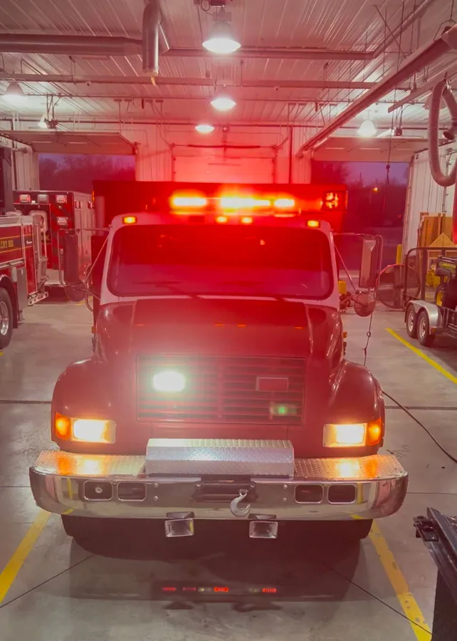 A red emergency apparatus in a station bay with its repaired roof light bar lit red, plus amber corner lights and a white scene light on the grille.