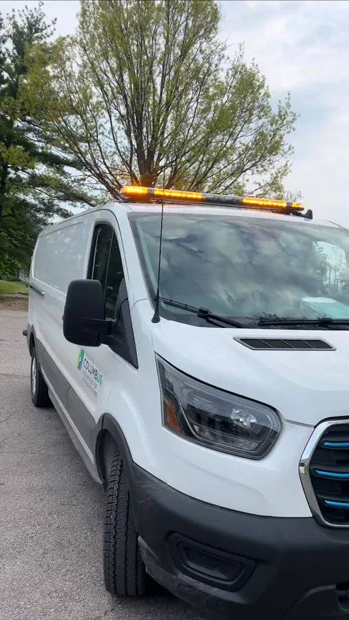 A white cargo van with a full-size amber LED light bar mounted across its roof rack, parked on asphalt in front of trees.
