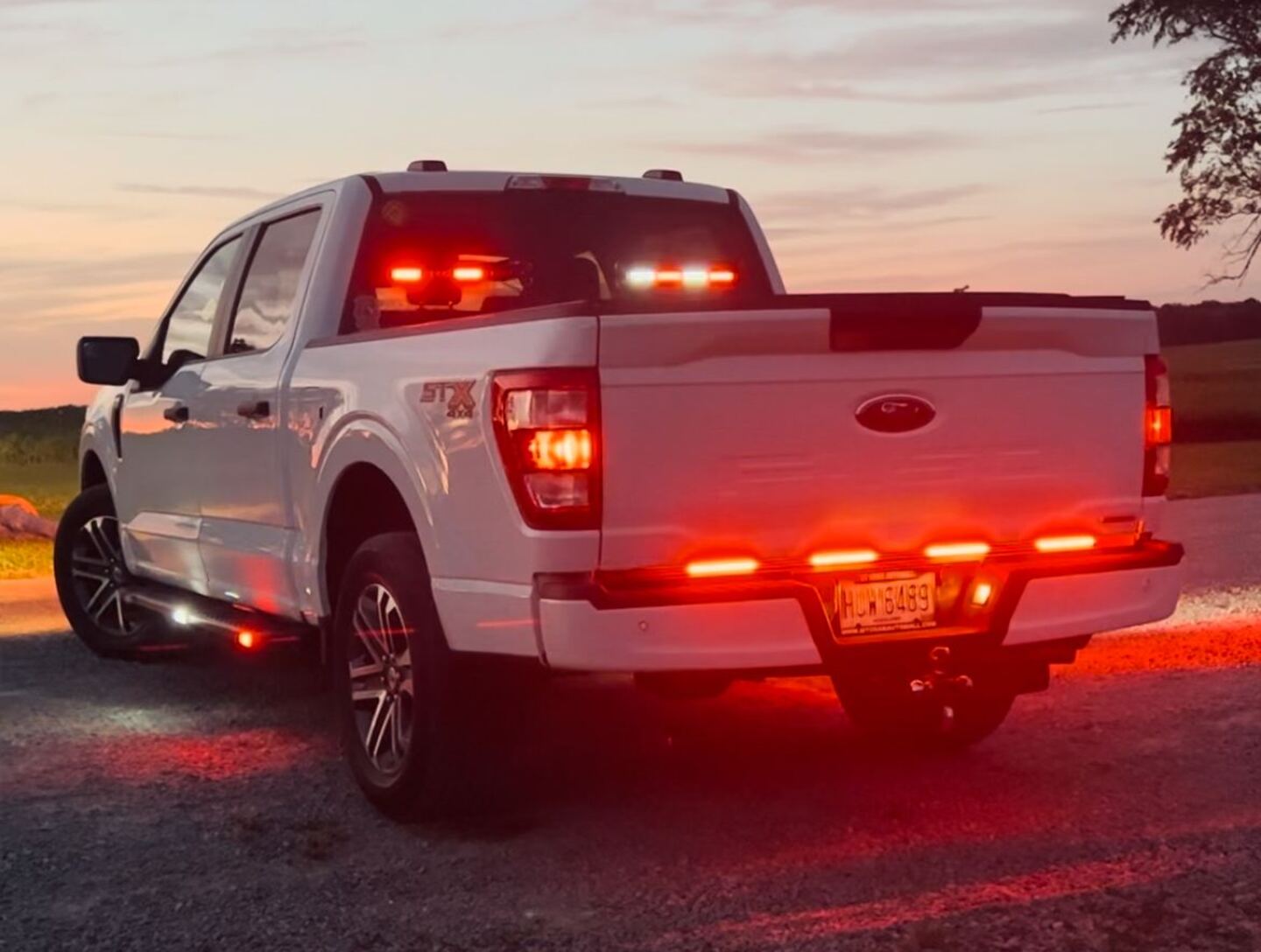 A white Ford F-150 at dusk with red LED warning lights lit across the cab roof, bed rail and lower bumper, throwing red light onto the gravel beneath it.