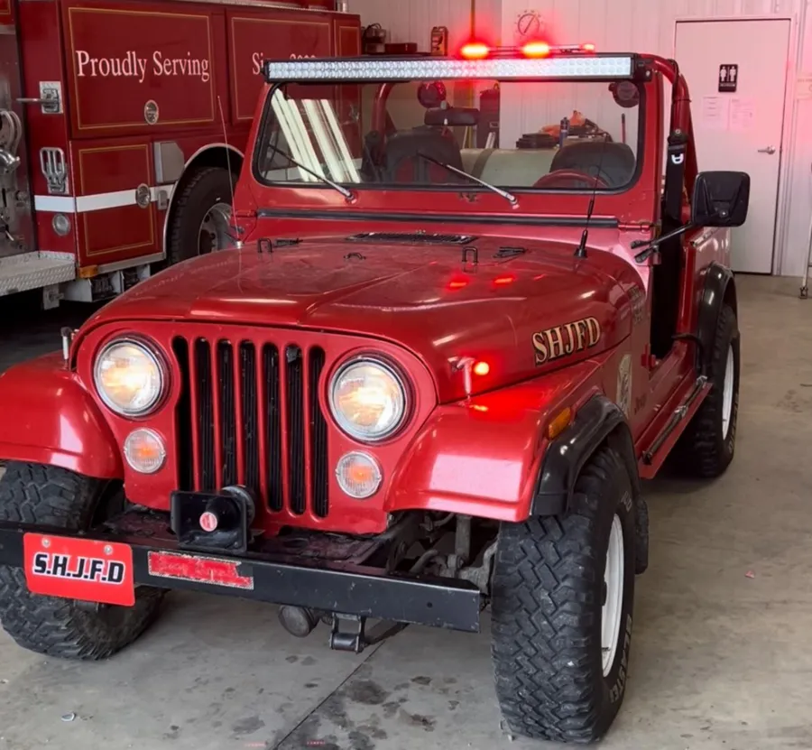 A red open-top brush unit inside a fire bay, carrying a long white LED light bar across the windscreen frame with red warning lights lit beside it and on the front fender.
