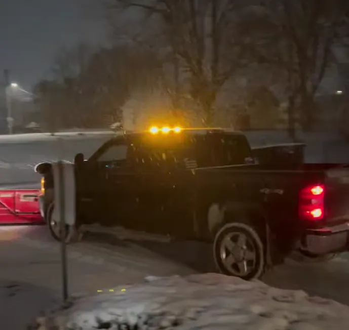 A dark pickup with a plow, running amber warning lights across its cab roof in falling snow at night on a snow-covered street.