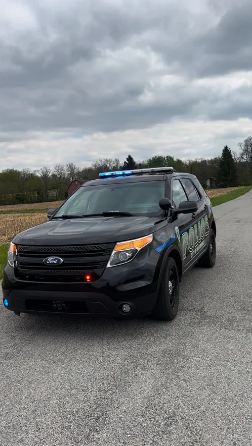 The same black police SUV from the front left on a rural road under overcast sky, roof bar unlit, a blue corner light and a red grille light showing.