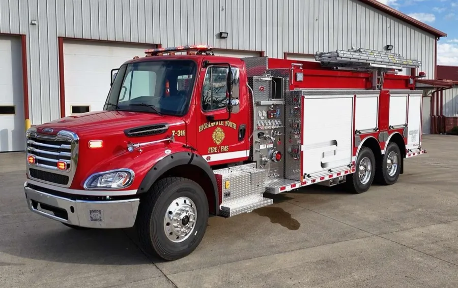 A red pumper-tanker with a chrome grille, hose bed and ground ladders parked on concrete outside a metal apparatus building.