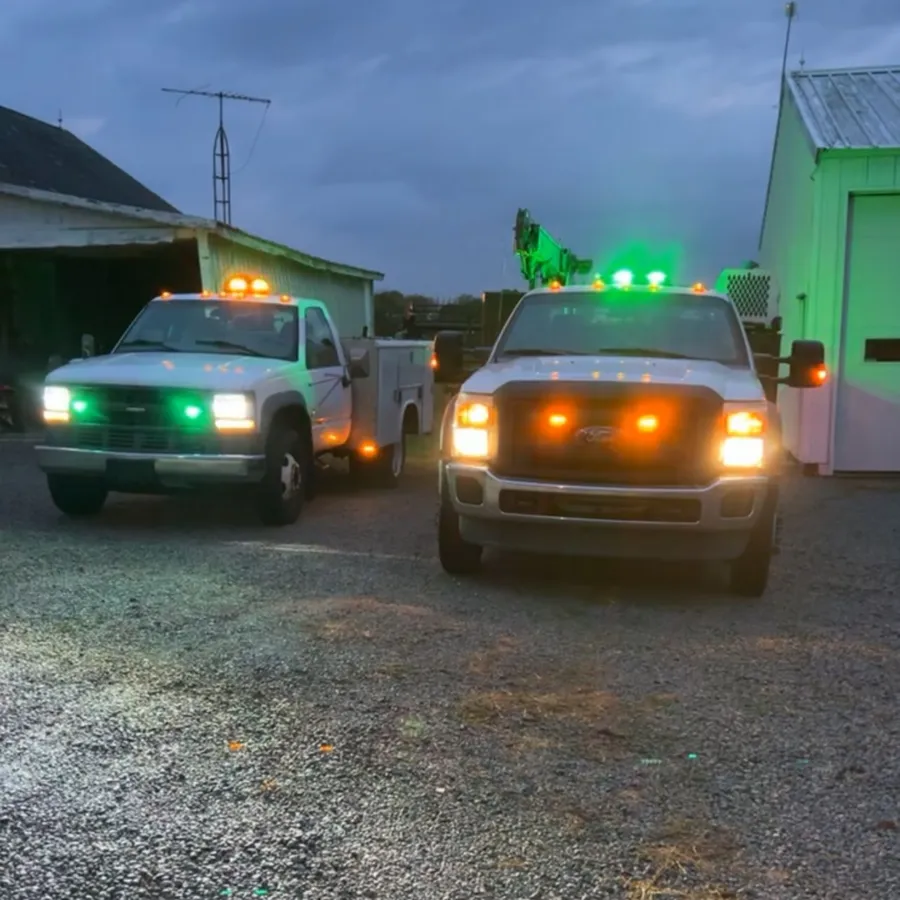 Two white service-bed trucks parked on gravel at dusk, both running amber roof and grille warning lights with green lights lit at the headlamps and roof.