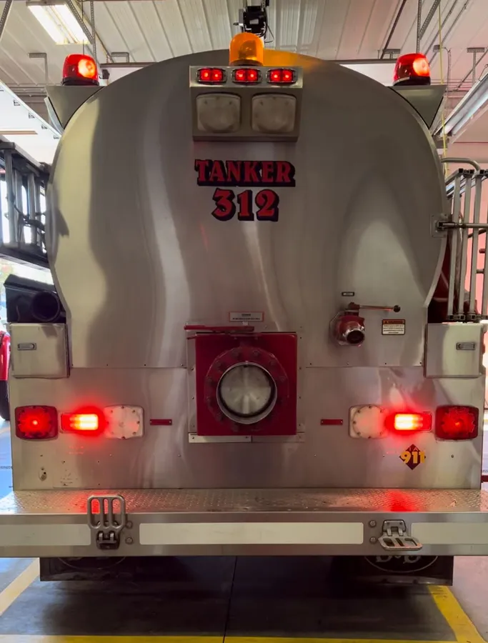 The polished aluminium rear of a fire tanker lettered Tanker 312, with red warning lights lit at the tank top and at both lower corners inside a station bay.
