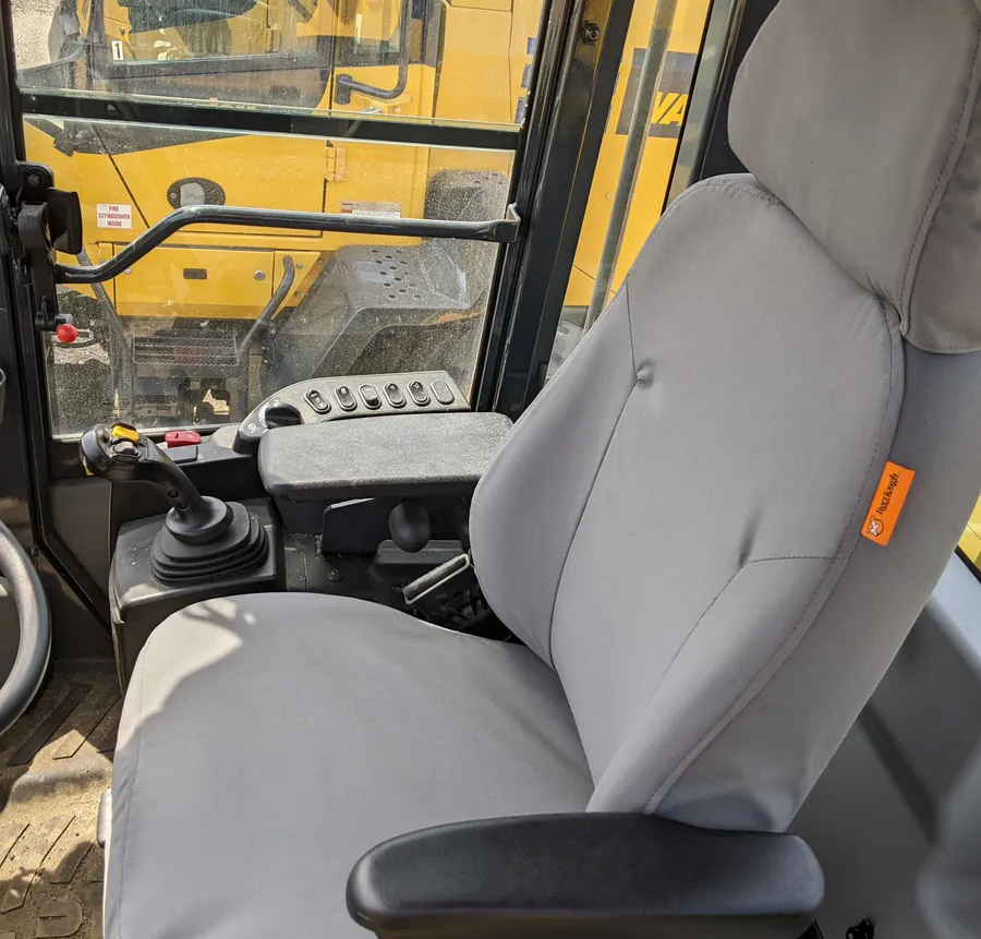 A grey TigerTough cover on the operator seat of a heavy equipment cab, the controls and a yellow machine beyond.
