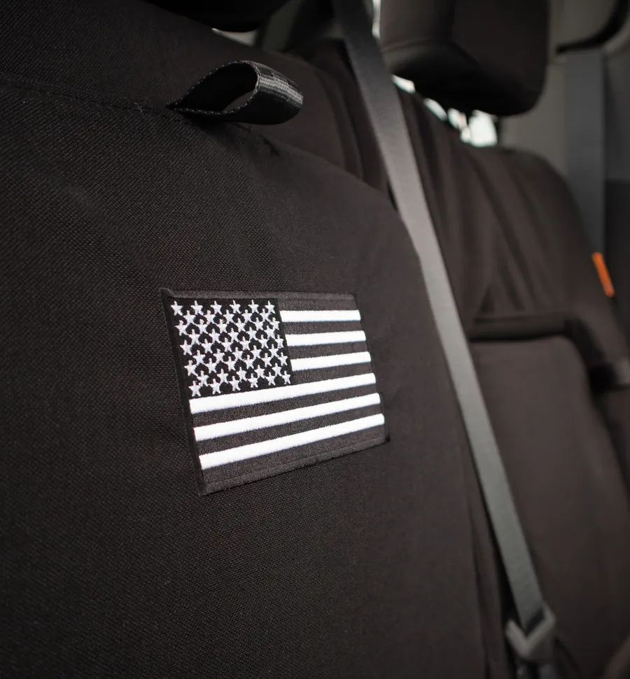 An embroidered American flag on the back of a black seat cover, the seat belt crossing beside it.