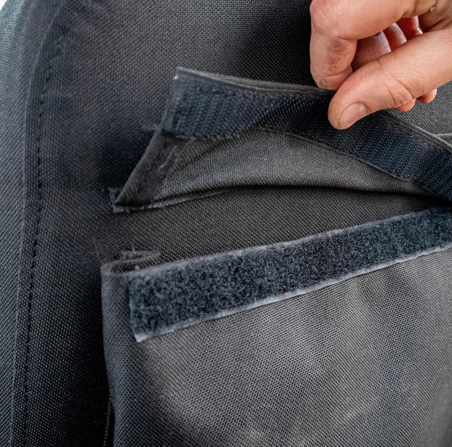 A hand lifting the hook-and-loop flap of a seatback pocket on a grey cover.