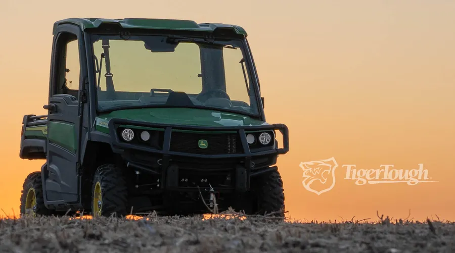 A green John Deere Gator side-by-side parked on a harvested field at sunset, the TigerTough logo in the corner.