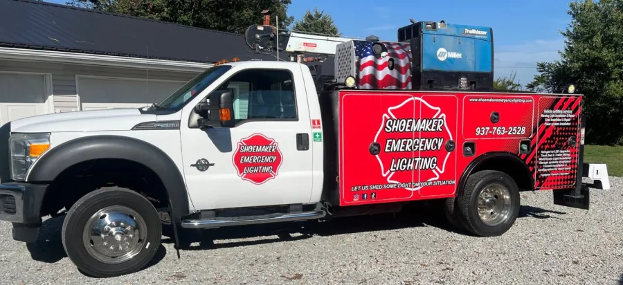 A white Ford service truck with a red Shoemaker Emergency Lighting wrap along the bed, carrying a welder and an American flag, parked on gravel beside a building.